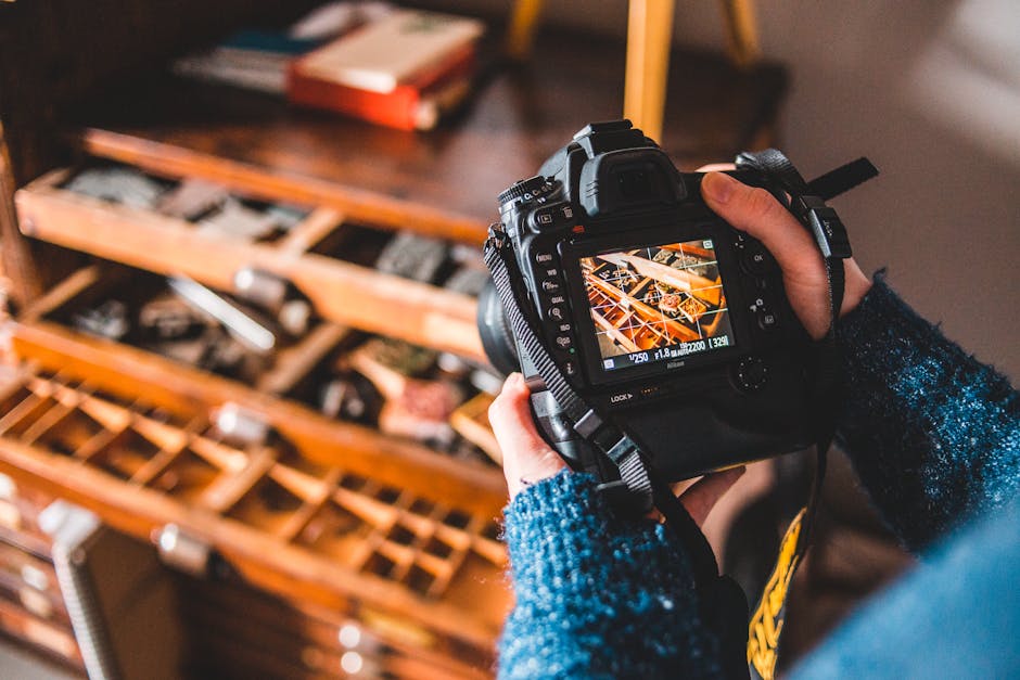 From above crop anonymous female photographer in blue sweater taking pictures on photo camera while standing near old cabinet with drawers filled with craft instruments